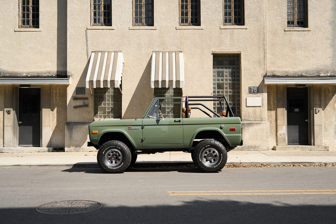 Classic Ford Bronco in Boxwood Green side profile parked on a city street, restored vintage Bronco with open top, roll cage, off-road tires, and custom suspension by Classic Ford Broncos.