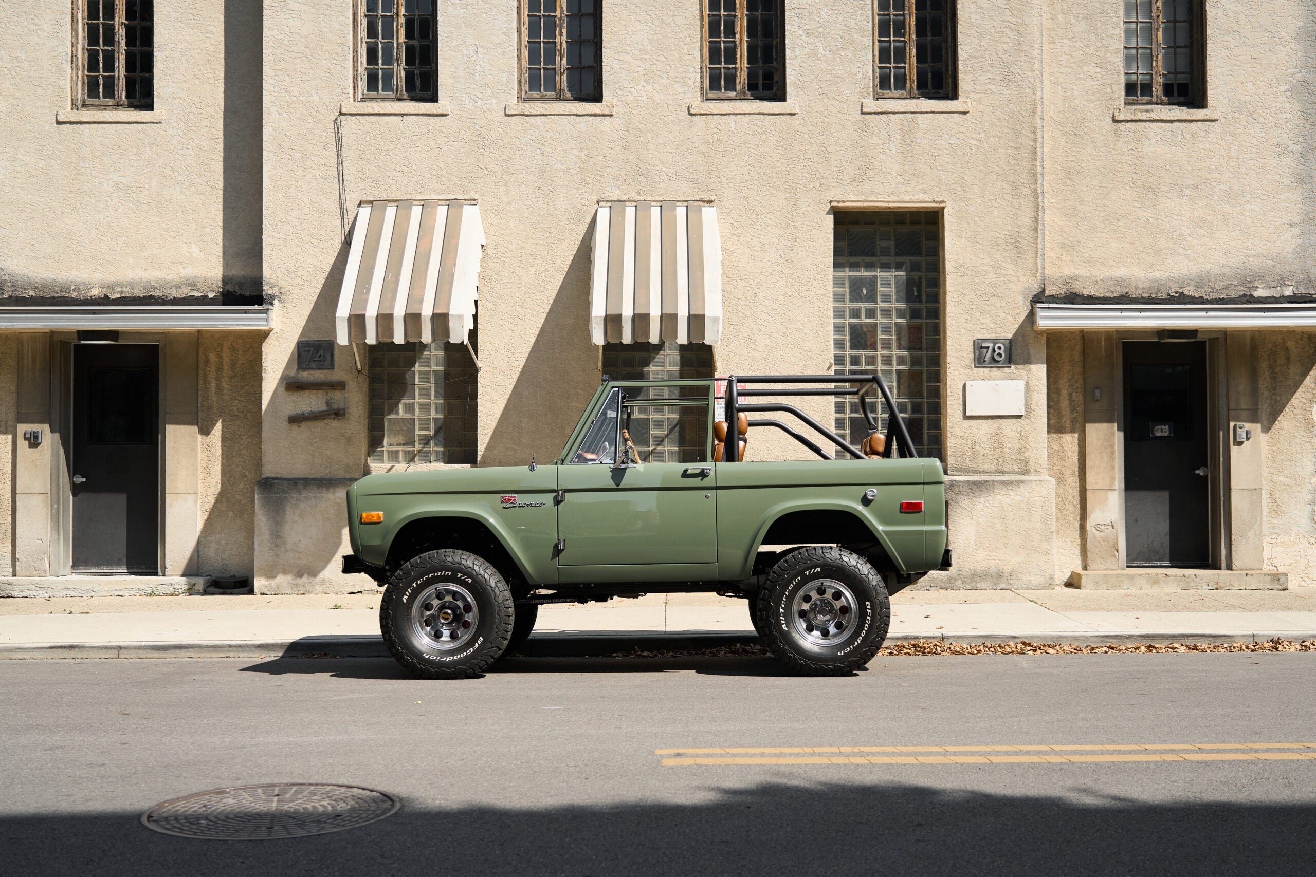 Classic Ford Bronco in Boxwood Green side profile parked on a city street, restored vintage Bronco with open top, roll cage, off-road tires, and custom suspension by Classic Ford Broncos.
