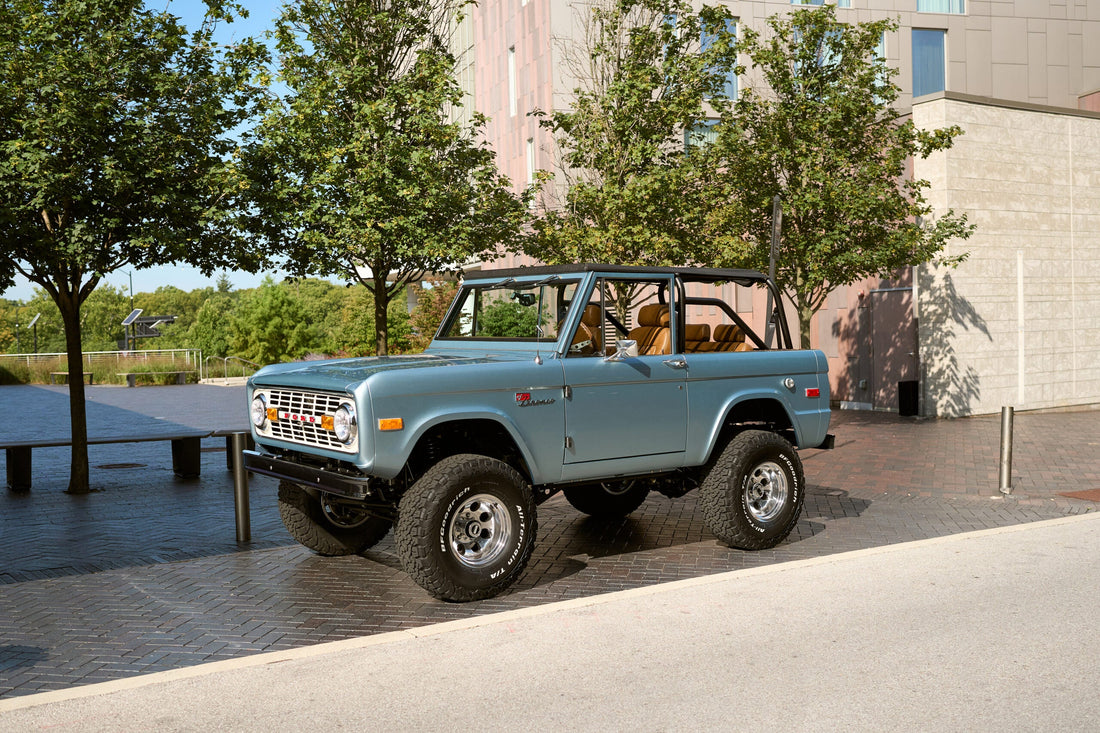 Brittany Blue 1966 Ford Bronco with Whiskey leather interior, parked curbside in an urban setting. Vintage Bronco restoration featuring Gen 3 Coyote 5.0L engine, lifted stance, and chrome accents.