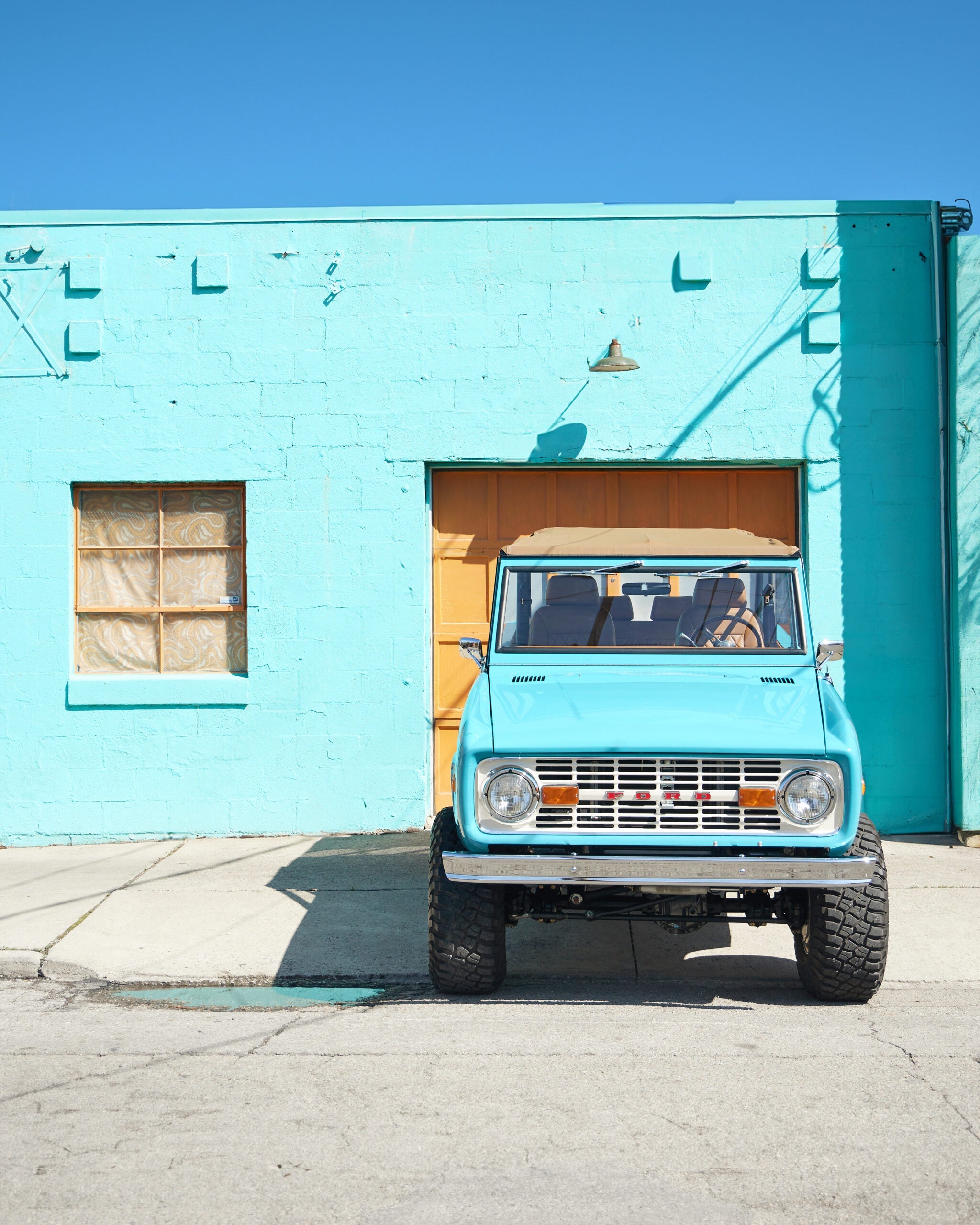 Front view of restored Classic Ford Bronco in Peacock Blue with tan soft top parked in front of a turquoise building, vintage early Bronco featuring lifted suspension, off-road tires, and custom restoration by Classic Ford Broncos.