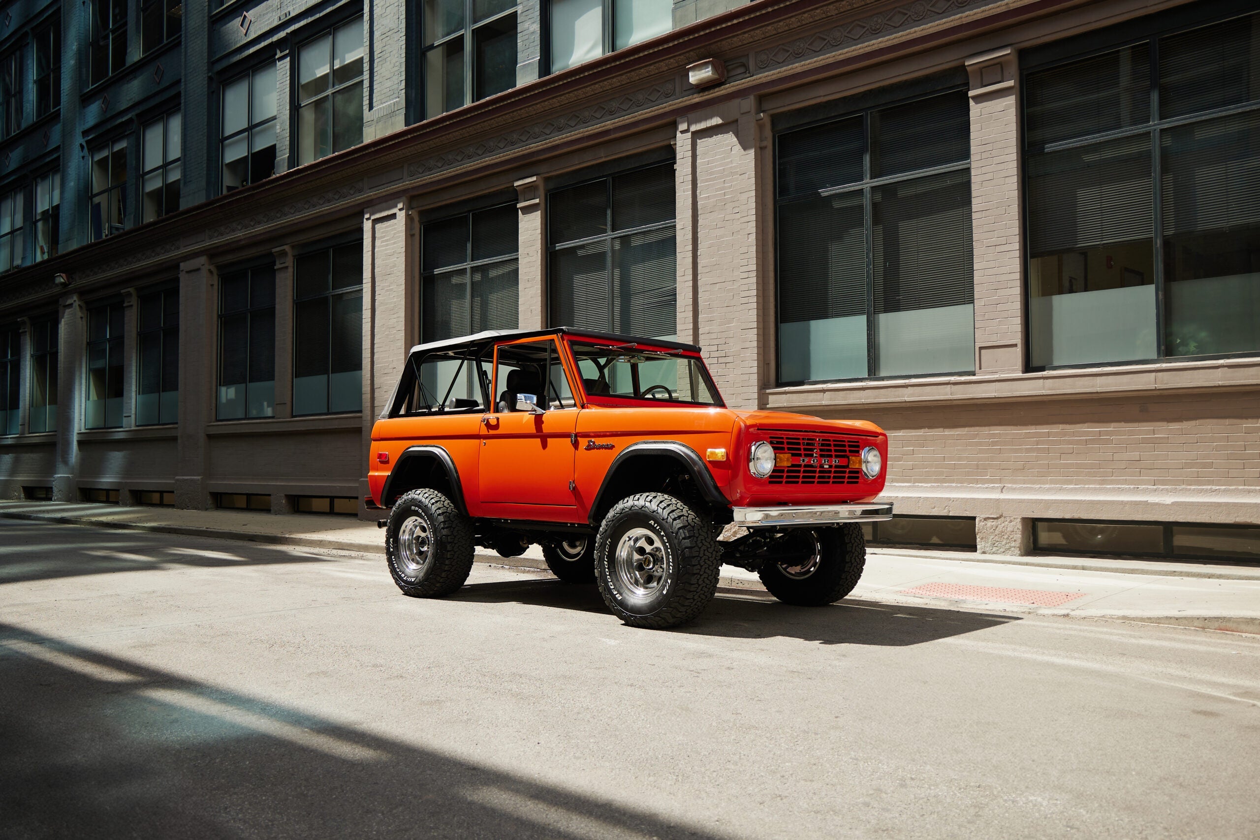 Classic Ford Bronco in bright orange parked on a city street, featuring lifted suspension, off-road tires, chrome wheels, and vintage styling with removable hardtop.