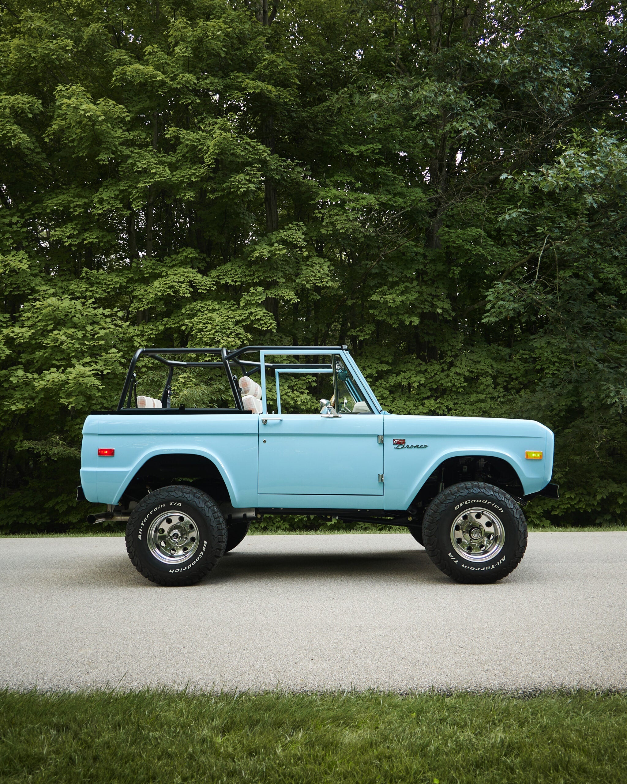 A fully restored 1970 Classic Ford Bronco named “Marblehead” is parked on a tree-lined road, featuring a lifted stance, roll cage, and open cabin with custom interior.