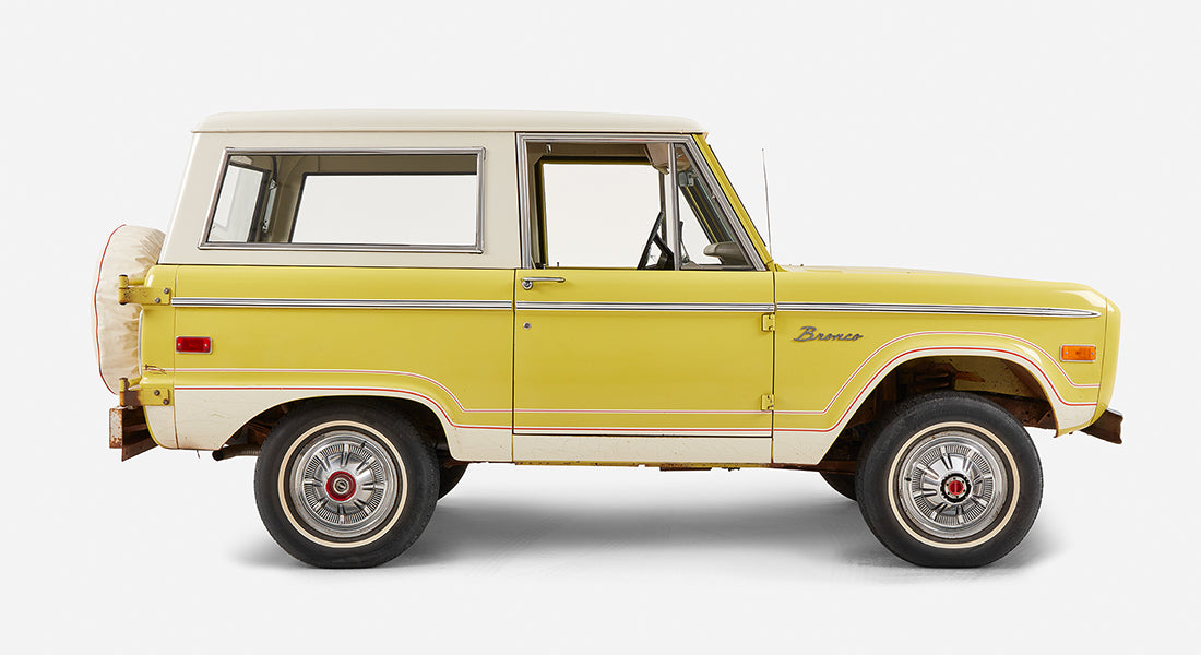 Side profile of yellow Classic Ford Bronco with white roof on a white background