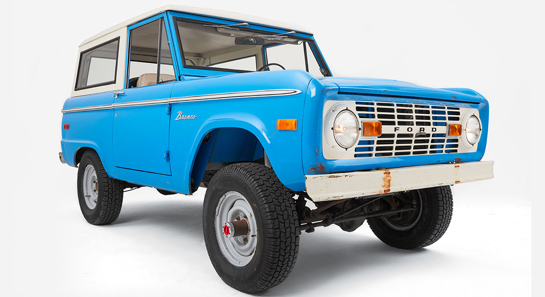 Blue Classic Ford Bronco with white roof and beige interior on white background, angled front view
