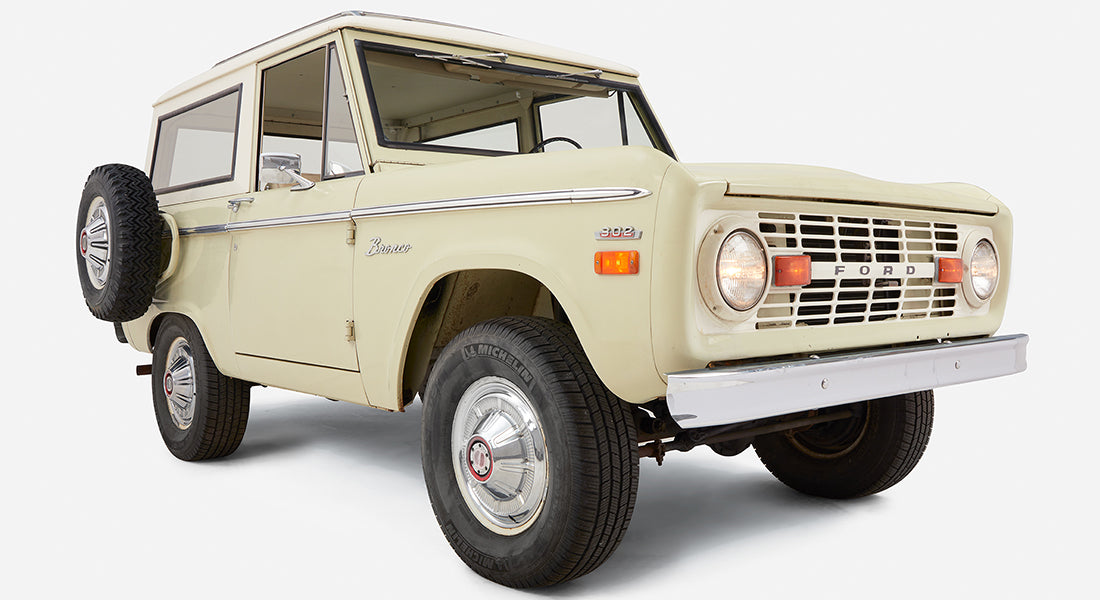 Light sage green Classic Ford Bronco with white roof, spare tire on rear, angled front view