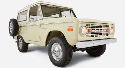 Light sage green Classic Ford Bronco with white roof, spare tire on rear, angled front view