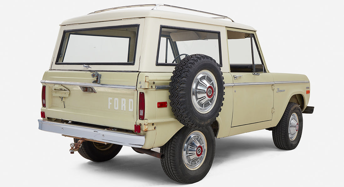Rear side view of a pale yellow Classic Ford Bronco with white top and full-size spare tire on the back