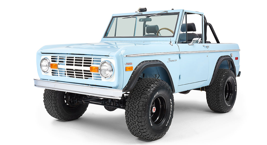 Side front view of light blue Classic Ford Bronco with black wheels and raised suspension on white background