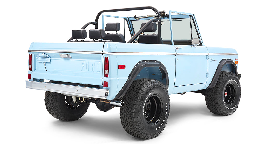 Rear three-quarter view of light blue Classic Ford Bronco with black wheels and roll bars on white background