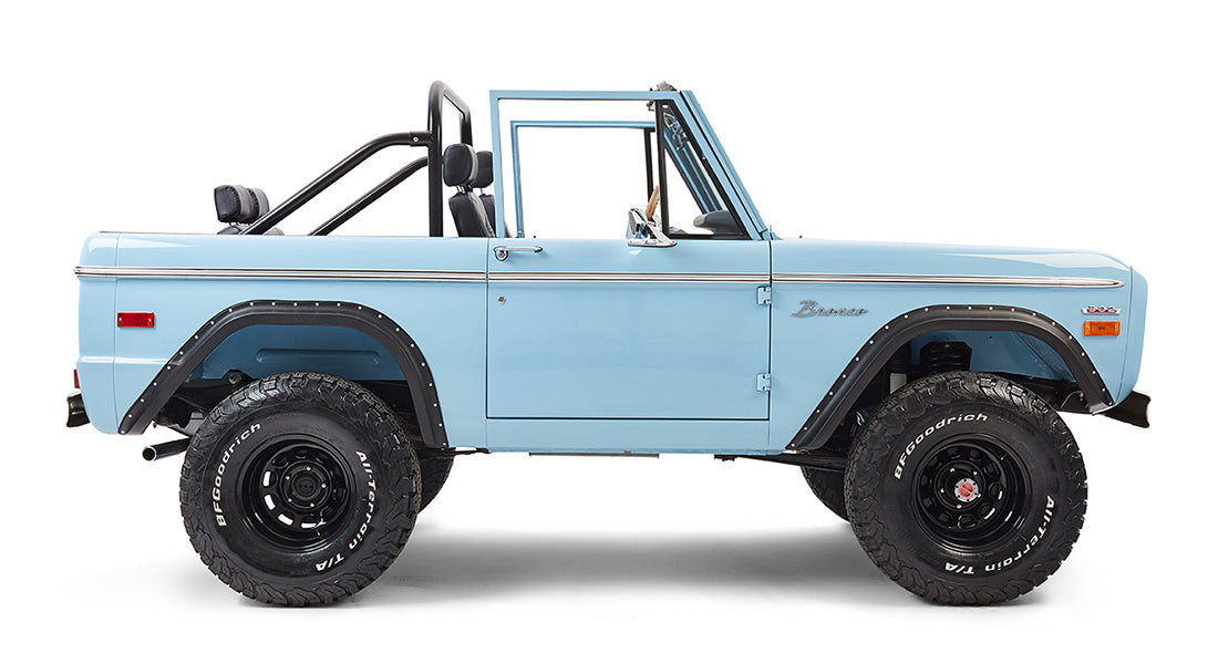 Classic Ford Bronco in baby blue with driver-side door visible, off-road tires, and roll bar.