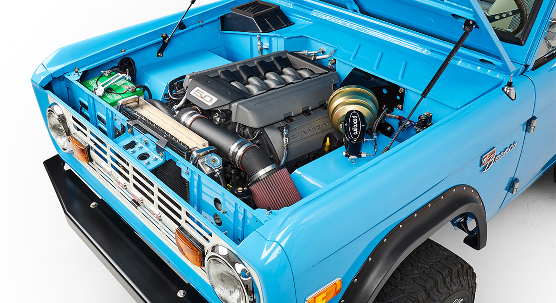 Angled front view of blue Classic Ford Bronco with raised hood showing engine and front fender