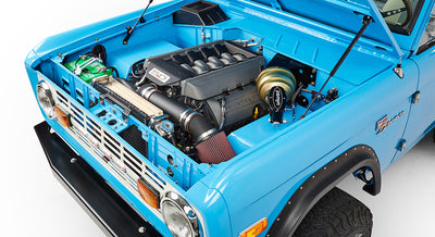Angled front view of blue Classic Ford Bronco with raised hood showing engine and front fender