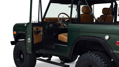 Open driver side door of Classic Ford Bronco showing tan leather seats and interior with visible roll cage