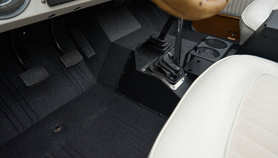 Black pedals and gear levers on black floor mat inside Classic Ford Bronco with white seats