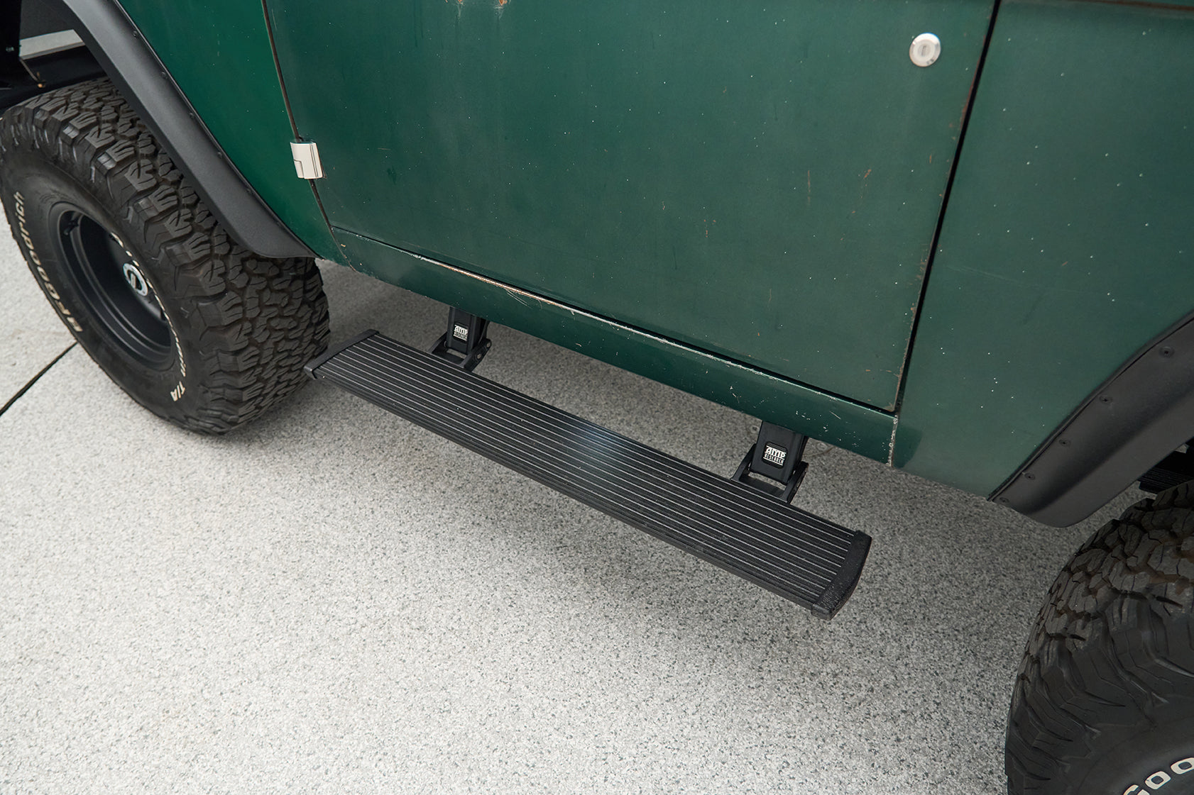 Black running board mounted below green vehicle door with large tire nearby