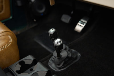 Close-up of two black gear shift levers and a tan leather car seat interior section.