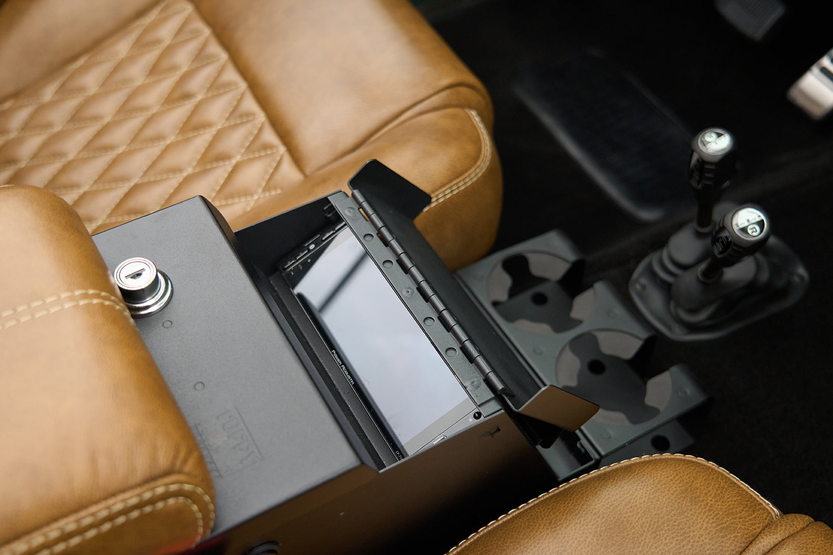 Car center console with brown leather seats, cup holders, key lock, and an open compartment showing a screen inside.