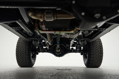 Underside view of a vehicle showing suspension, tires, and exhaust components on a gray floor.