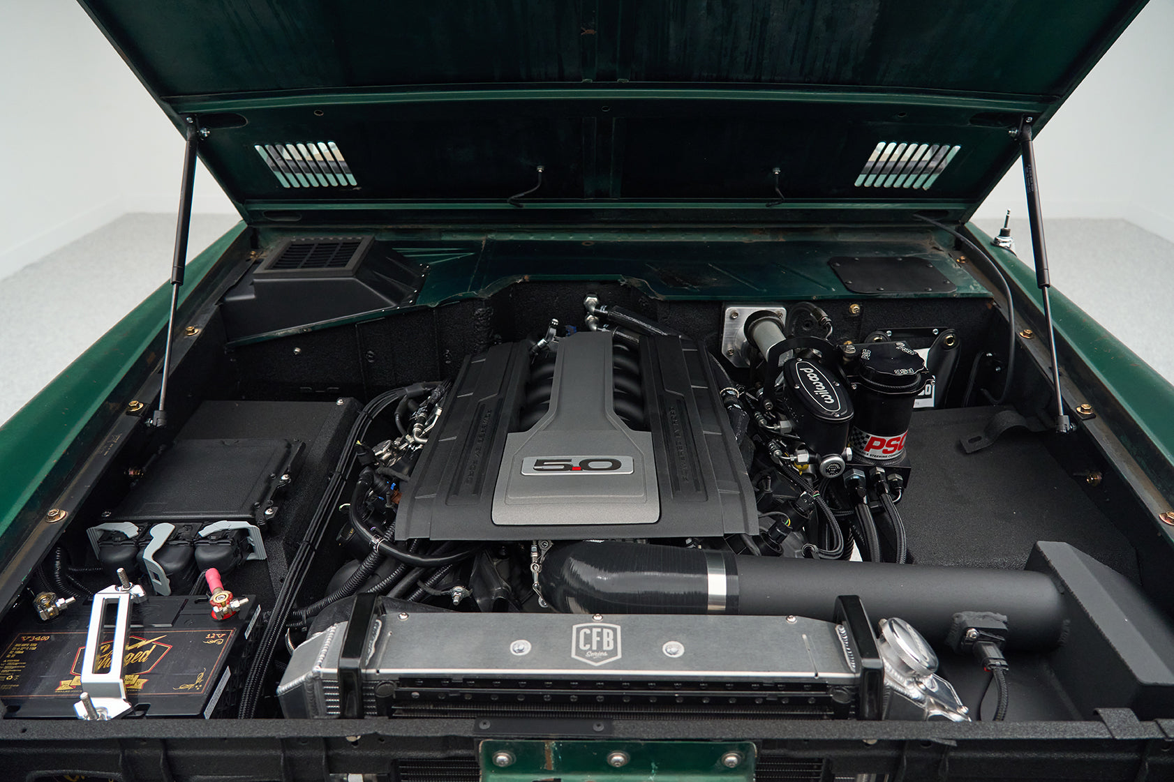 Engine bay of a Classic Ford Bronco showing a clean, modern 5.0-liter engine with visible components and battery.
