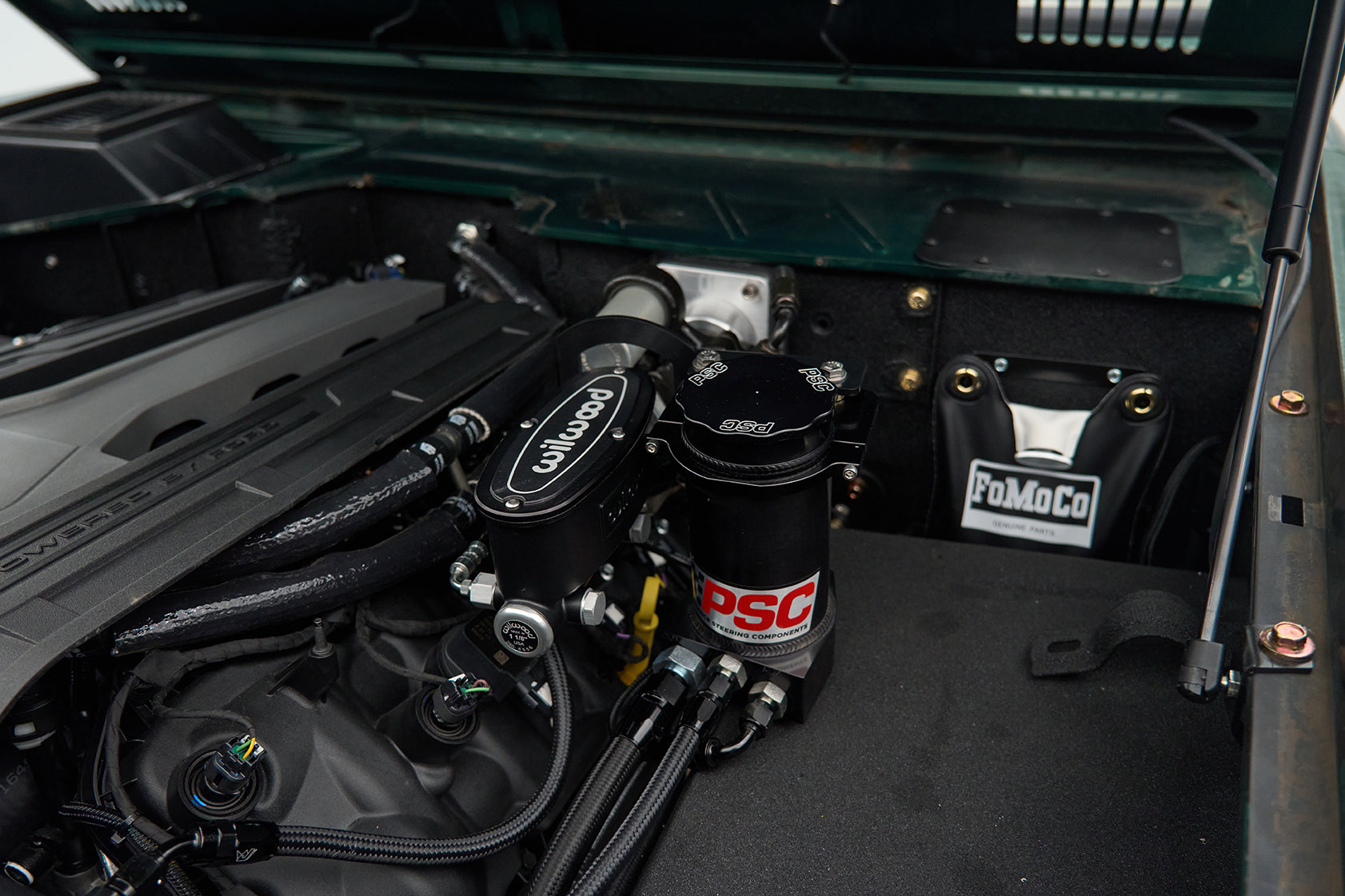 Engine compartment showing Wilwood and PSC power steering components with a FoMoCo parts bag in the background.