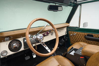 Wood-rimmed steering wheel and tan dashboard with Classic Ford Bronco gauges visible inside vehicle.