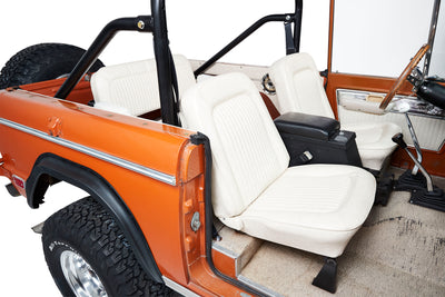 Classic Ford Bronco interior with white seats, orange body, roll bar, and off-road tires.