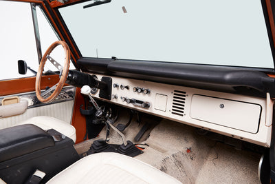 Interior dashboard and steering wheel of a Classic Ford Bronco with manual gear shifters and white seats.