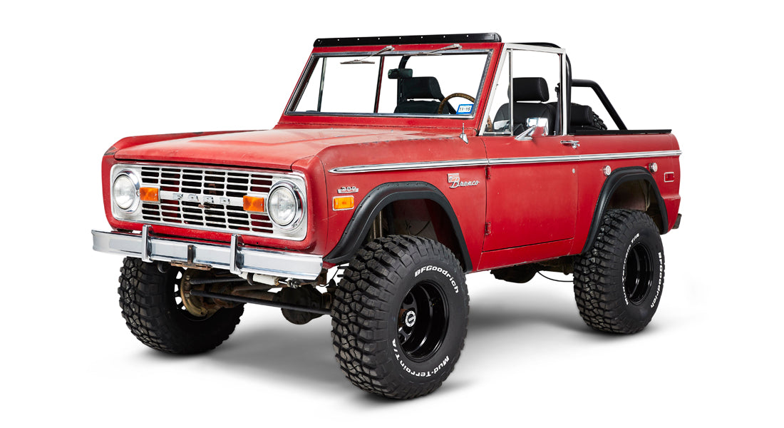 Front three-quarter view of red Classic Ford Bronco with open roof and large off-road tires on white background.