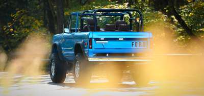 Classic Ford Bronco rear view in blue with brown seats and roll cage, surrounded by blurred foliage.