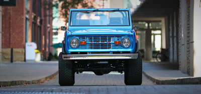 Classic Ford Bronco front view in blue parked on a cobblestone street between buildings.