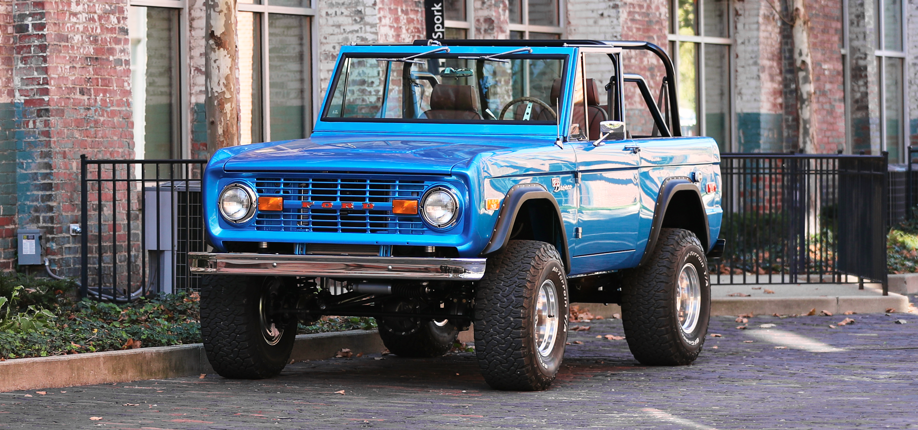 Classic Ford Bronco angled front view in blue, parked on a cobblestone street with buildings in background.