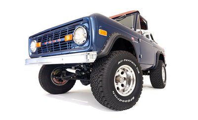 Low-angle front view of blue Classic Ford Bronco with raised suspension and all-terrain tires on white background