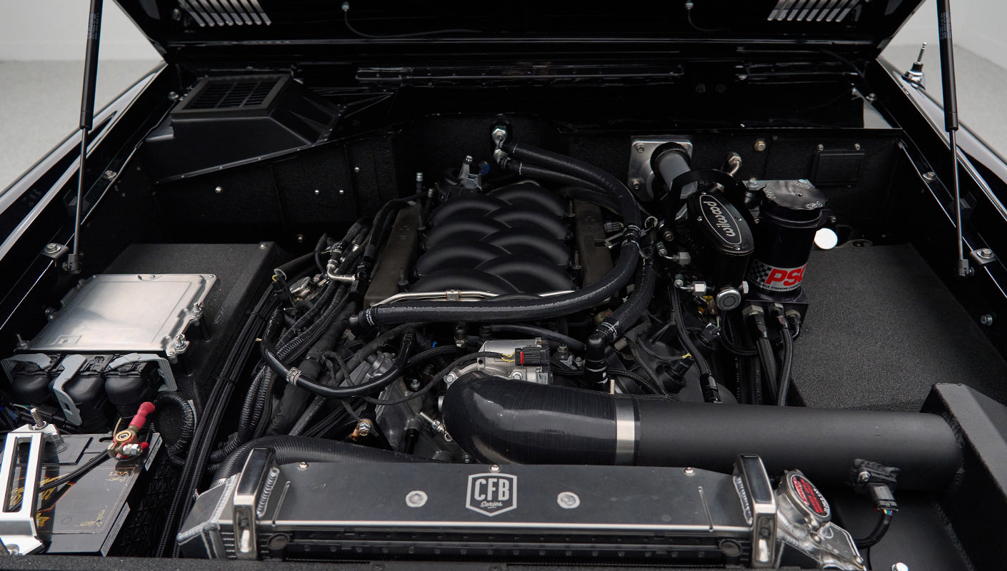 Engine bay of Classic Ford Bronco with black intake tubes, battery, and various fluid reservoirs