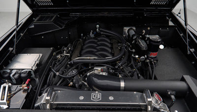 Engine bay of Classic Ford Bronco with black intake tubes, battery, and various fluid reservoirs