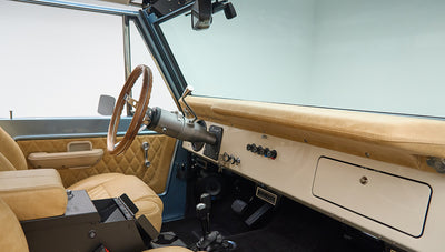 Classic Ford Bronco interior showing beige dashboard, black floor, and wooden steering wheel.
