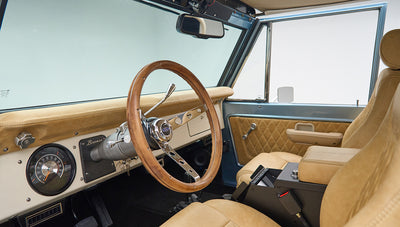 Classic Ford Bronco driver seat and dashboard with wooden steering wheel and beige upholstery.