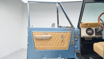 Classic Ford Bronco driver door with beige diamond-stitched door panel and blue exterior.