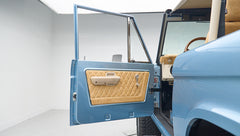 Open driver-side door showing blue exterior and beige diamond-stitched door panel of Classic Ford Bronco.