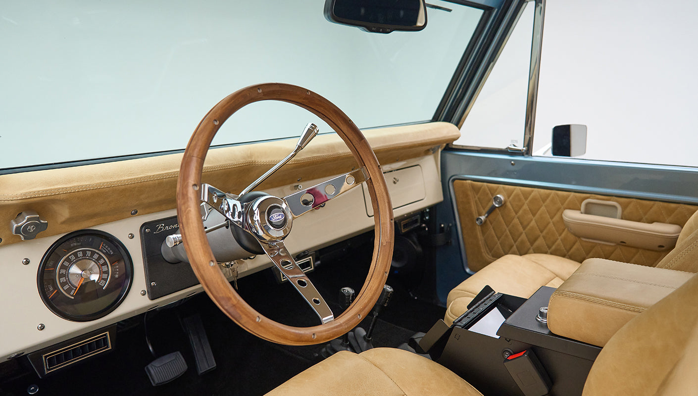 Classic Ford Bronco driver seat and dashboard with wooden steering wheel and beige upholstery.