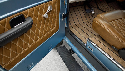Open door of Classic Ford Bronco showing blue exterior and quilted brown leather door panel with manual window crank