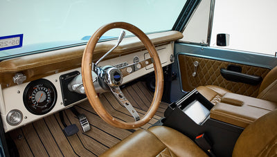 Driver-side interior of Classic Ford Bronco showing wooden steering wheel, brown leather seats, and dashboard