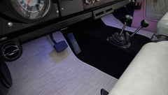 Foot pedals and two gear levers on black carpet floor with beige and black surrounding in a Classic Ford Bronco