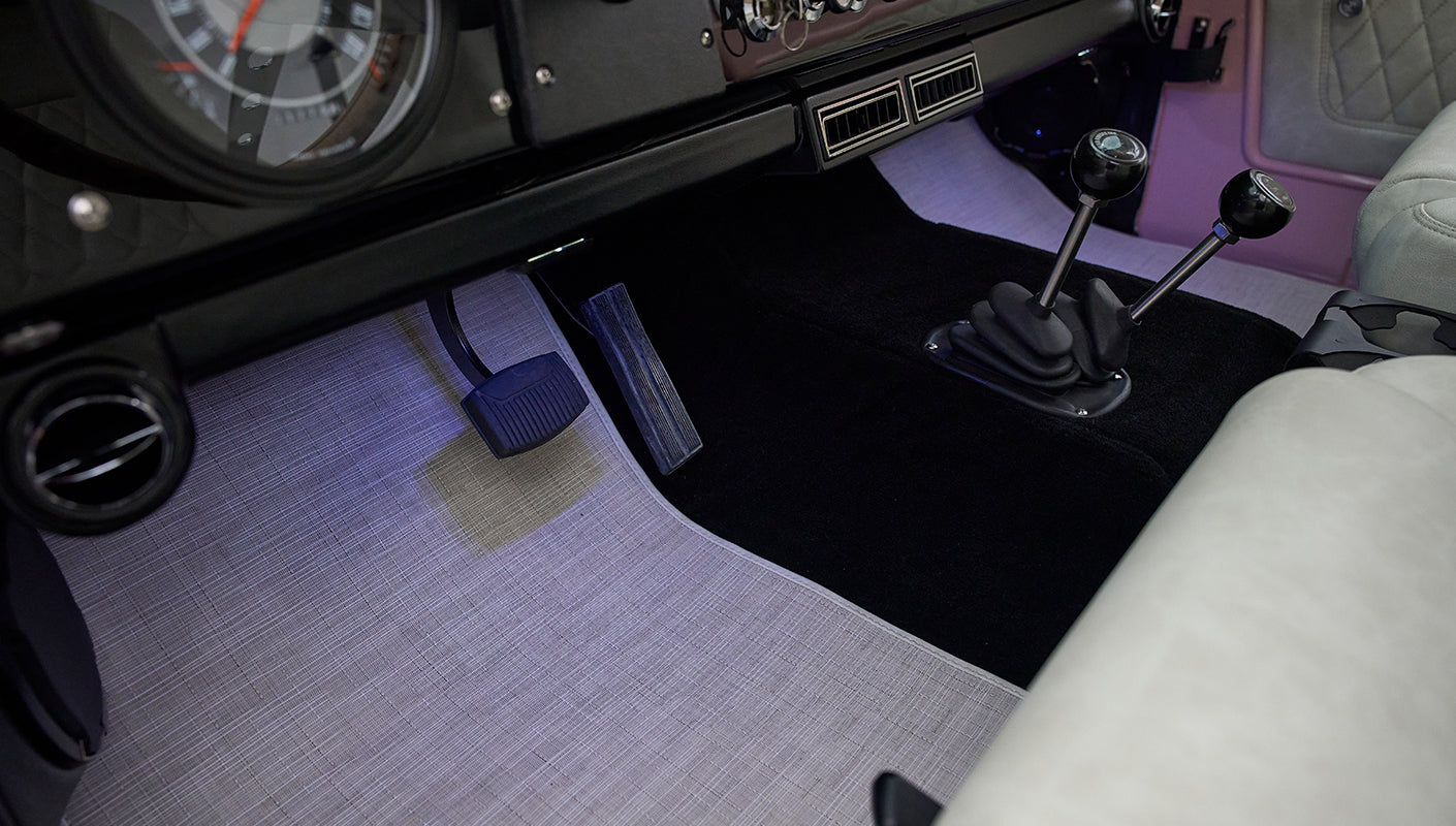 Foot pedals and two gear levers on black carpet floor with beige and black surrounding in a Classic Ford Bronco