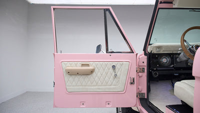 Open driver door of a pink Classic Ford Bronco showing quilted upholstery on the door panel and part of steering wheel.