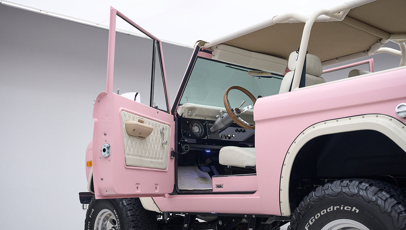 Side view of pink Classic Ford Bronco with open driver door displaying cream interior and wood-accented steering wheel.