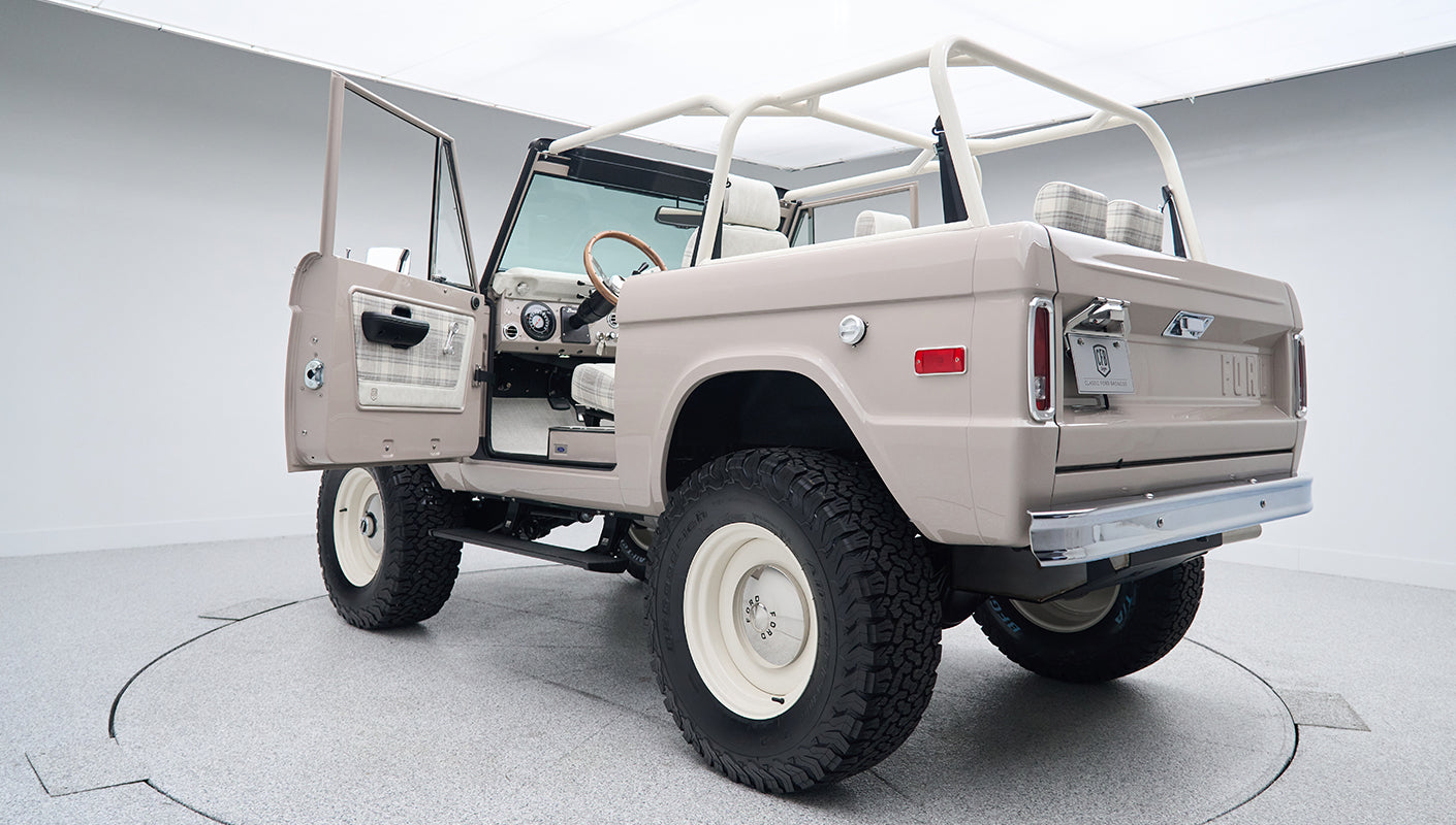 Rear exterior view of classic Ford Bronco with open driver door, white wheels, and beige body