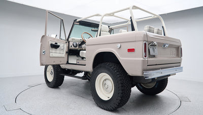Rear exterior view of classic Ford Bronco with open driver door, white wheels, and beige body
