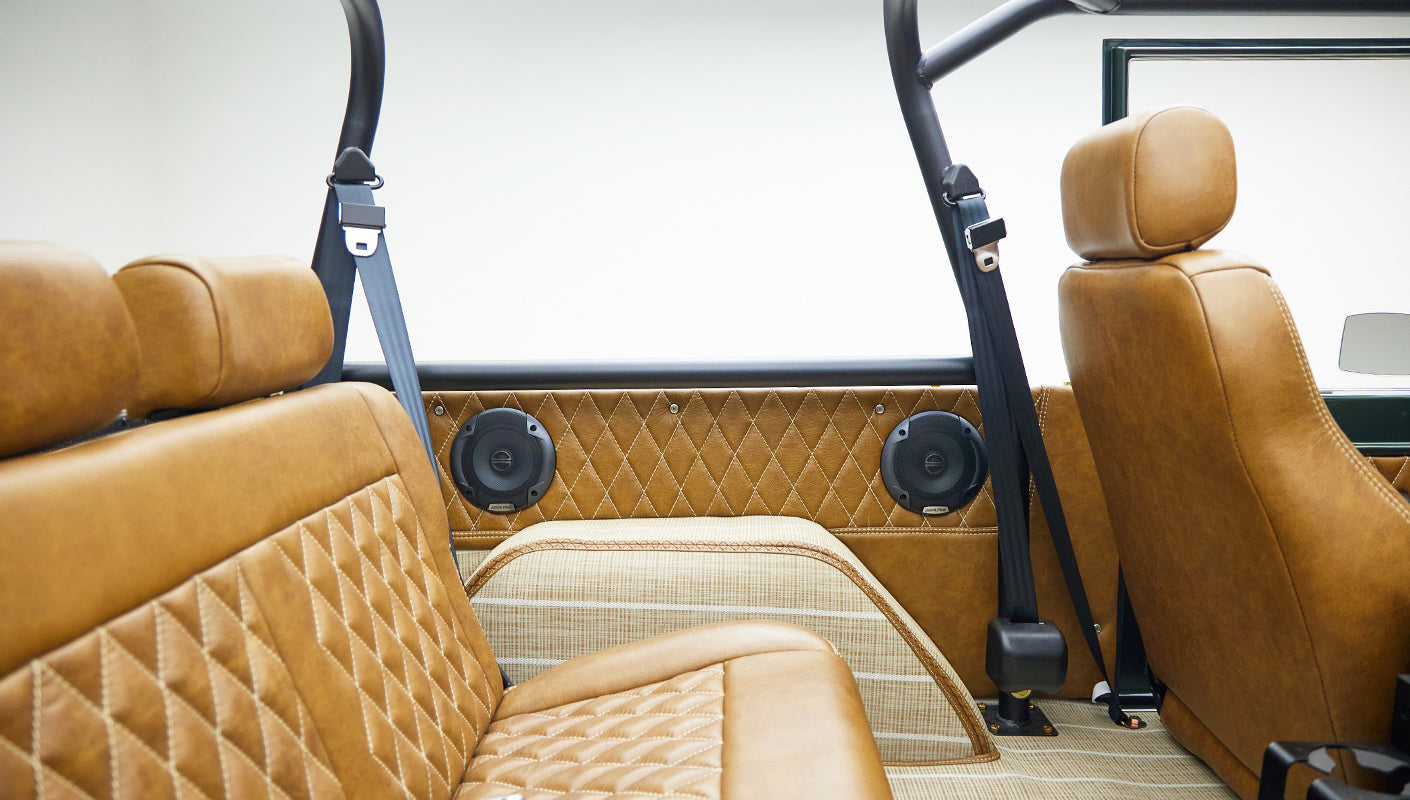 Interior of a Classic Ford Bronco with tan diamond-stitched seats, black seat belts, and rear speakers on the side panel.