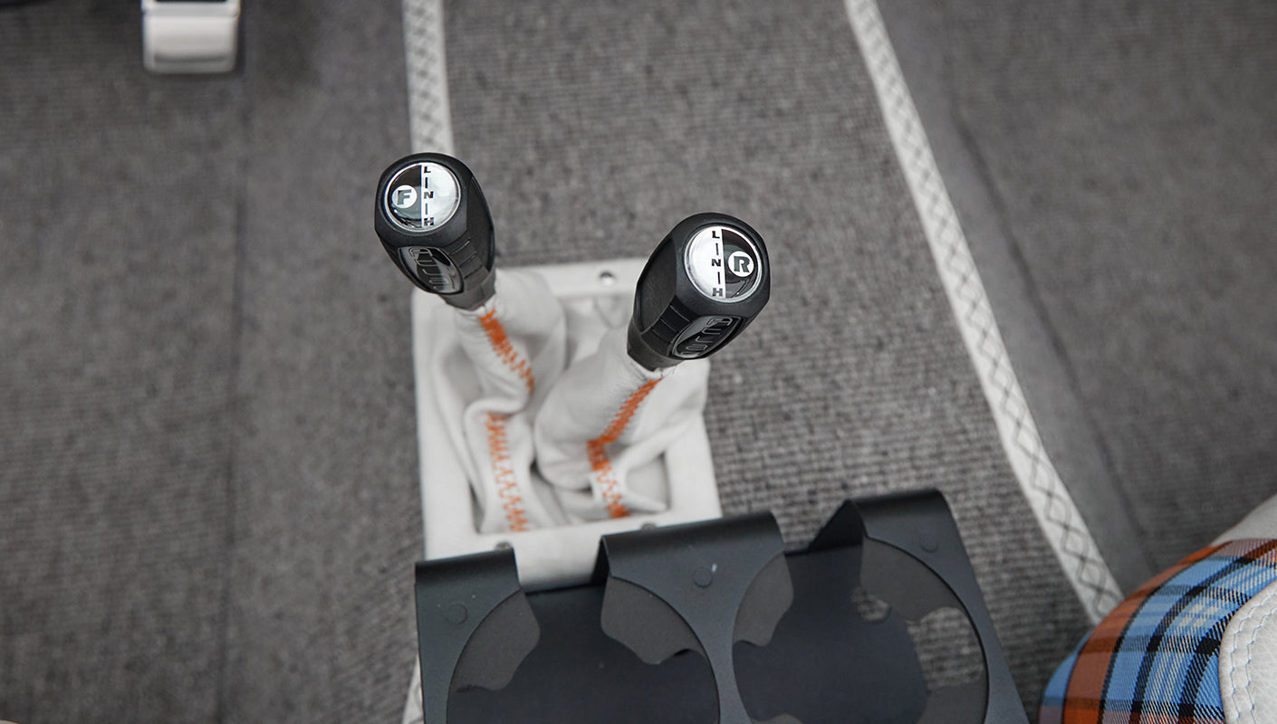 Console with two black gear shift knobs with white leather covers and orange stitching on gray carpet floor