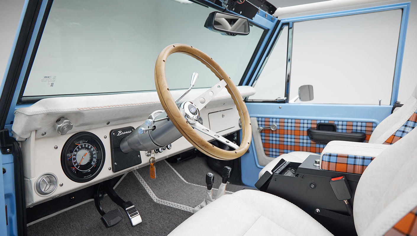 Interior of Classic Ford Bronco showing wooden steering wheel, white seats, gray flooring, and plaid door panel